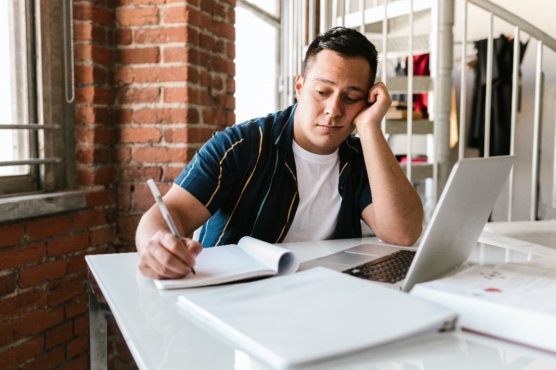 A young man looks at a notebook while using his laptop, clearly frustrated with his slow Internet speeds. 